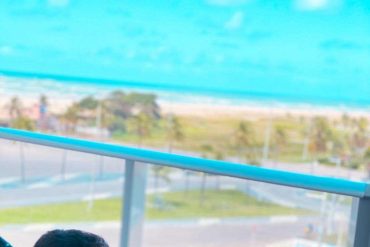 Homem sorridente usando laptop em um ambiente moderno de home office, com gráficos de crescimento e ícones de marketing digital ao redor, representando o Método TGR de Maycon Thiaillen.