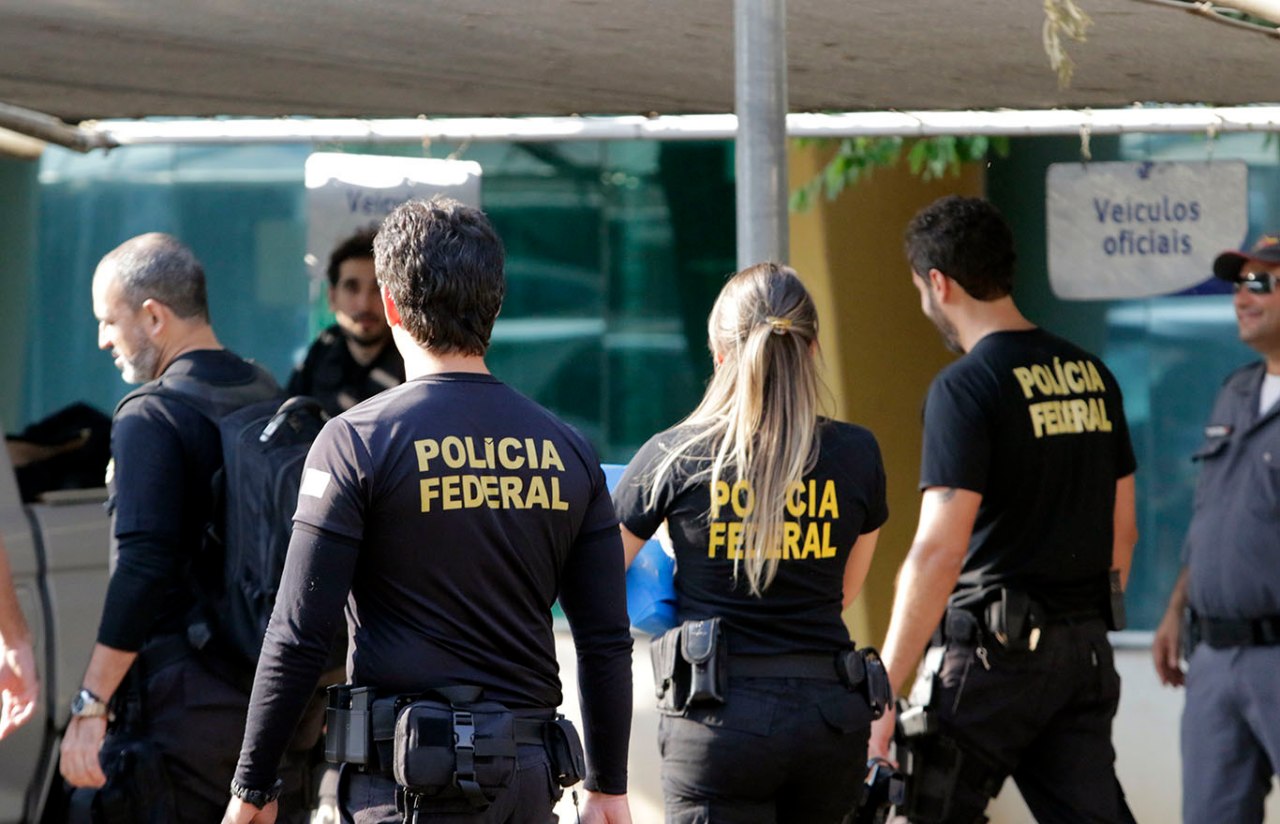 Homem sorridente em uniforme da Polícia Federal, segurando um livro de estudos com um escudo da PF ao fundo em um ambiente digital.